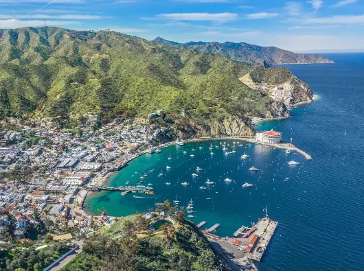 Sunlit Aerial Panorama of Avalon Harbor, Santa Catalina Island Aerial view of Avalon Harbor on Santa Catalina Island, showing the crescent-shaped bay dotted with boats, the iconic white Casino building on the waterfront, and the rugged green hills rising behind the coastal town.
